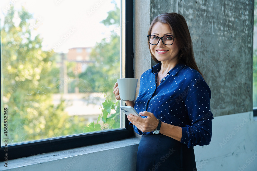 Fototapeta premium Middle aged beautiful businesswoman with smartphone, cup, near window