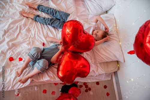 Two young children relaxing on a bed decorated with red heart balloons and rose petals, celebrating love and childhood happiness during Valentines Day