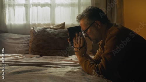 Side shot of mature man with grey hair holding bible book and rosary turning to God during morning prayer while kneeling with elbows on bed