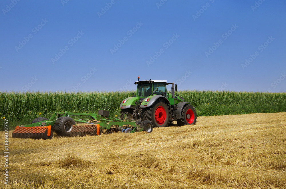 Fototapeta premium agricultural tractor working in the field after harvesting wheat next to a cornfield