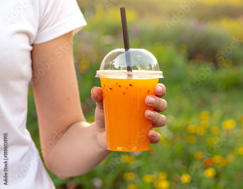 Person holds an orange beverage in a clear plastic cup with a straw, against a soft blurred background of flowers