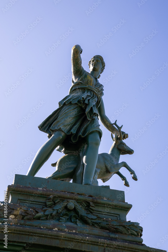 Fototapeta premium Classical statue of Diana the Huntress in Praza Galicia Fonte de Diana, Betanzos, Galicia, Spain. Historic stone fountain surrounded by lush greenery.