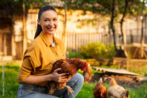 Portrait of happy young woman on private farm holding chicken and smiling at camera, posing on backyard farm, promoting organic poultry farming