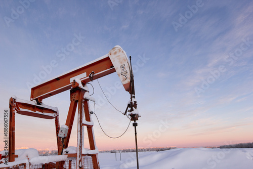 Oil pumpjack covered in snow against a soft pink winter sky at sunrise. Industrial energy production in rural Canada, cold season, oil and gas industry, prairie landscape.