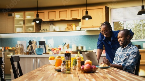 Tableau sur toile African American freelancer sitting at a kitchen table with a laptop, sending emails and explaining business tasks to his girlfriend