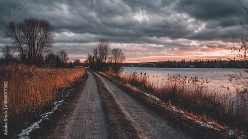 A winding dirt road leads towards a lake beneath a dramatic, cloudy sky during sunset