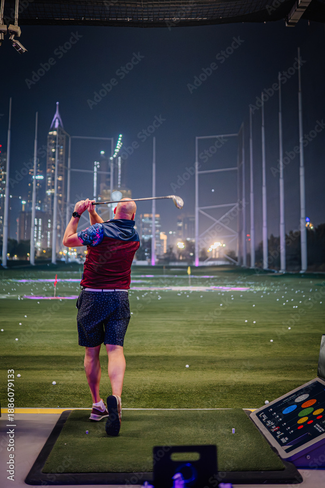 Fototapeta premium Man in colorful attire swings golf club at Topgolf Dubai, with city skyline illuminated at night in the background