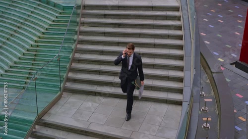 High angle tracking shot of young businessman in suit talking on smartphone while rushing down stairs carrying skateboard and laptop bag in modern urban environment