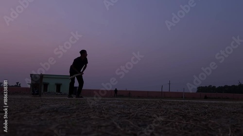 Silhouette of a unrecognized batsman standing near the pitch during calm sunset light.