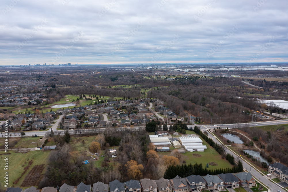 Fototapeta premium Suburban neighborhood in brampton, ontario with residential houses, a golf course, greenhouses, roads, and a visible toronto city skyline under an overcast sky
