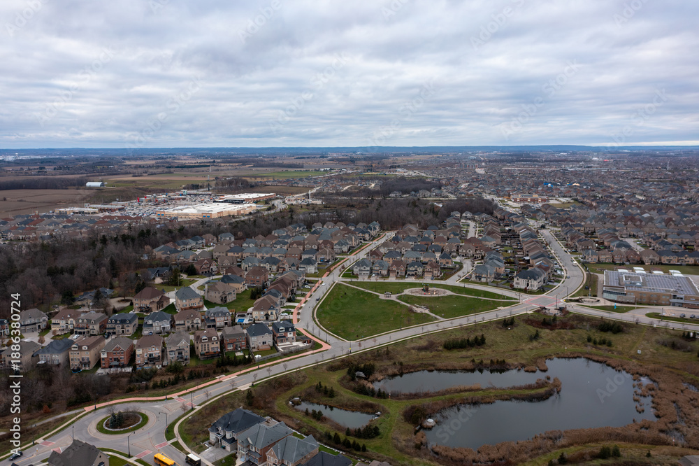 Fototapeta premium Aerial view showing suburban residential development with many houses. Streets. A roundabout. And retention ponds contrasting against open agricultural land and a commercial plaza in brampton. Ontario