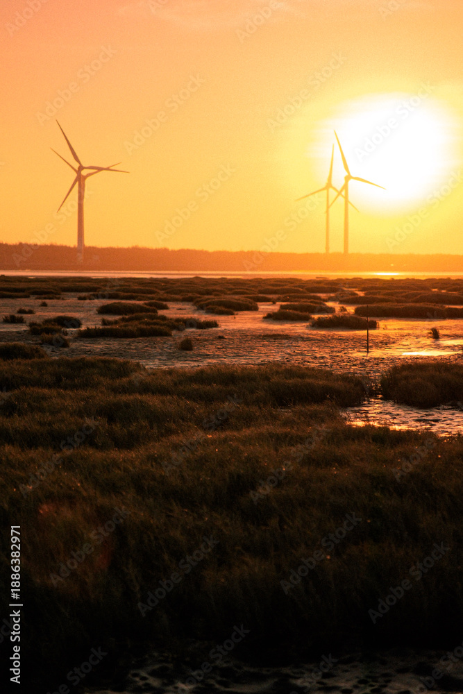 Obraz premium Sunset over coastal wetlands with wind turbines on the horizon, golden sky reflecting on tidal flats, calm water and silhouettes