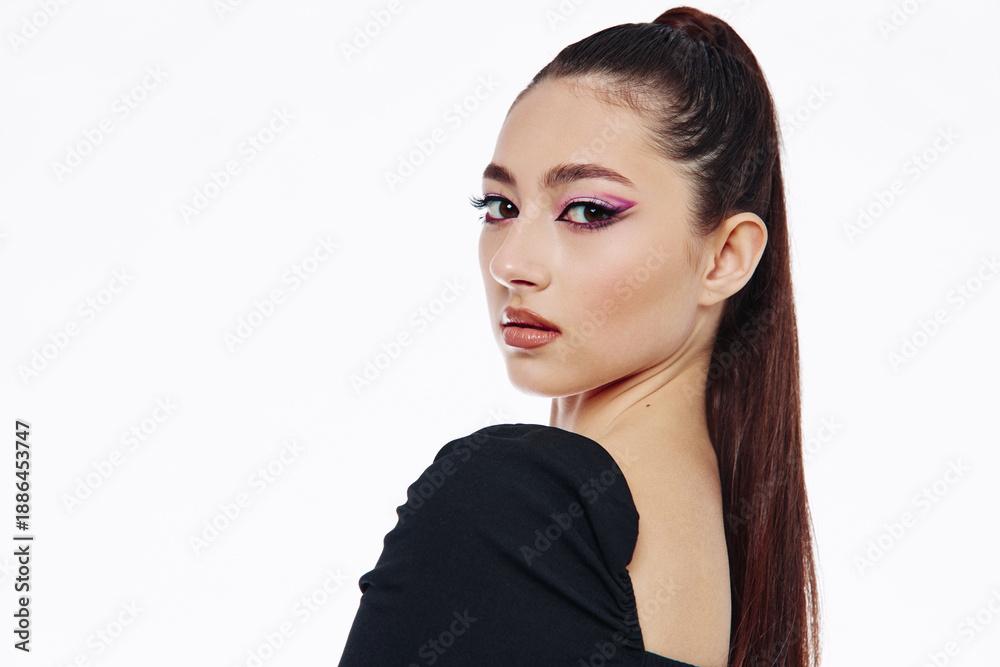 Naklejka premium Young woman with sleek dark ponytail and artistic pink eye makeup gazes confidently over her shoulder. Studio portrait on white background. Fashion and beauty creative concept.