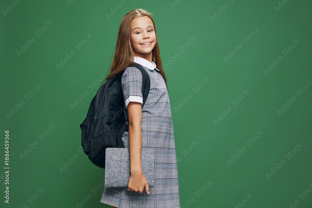 Fototapeta premium Girl, schoolchild and backpack standing with book for education at studio. Young student, smiling child holding notebook in casual dress with long hair on green background for learning concept.