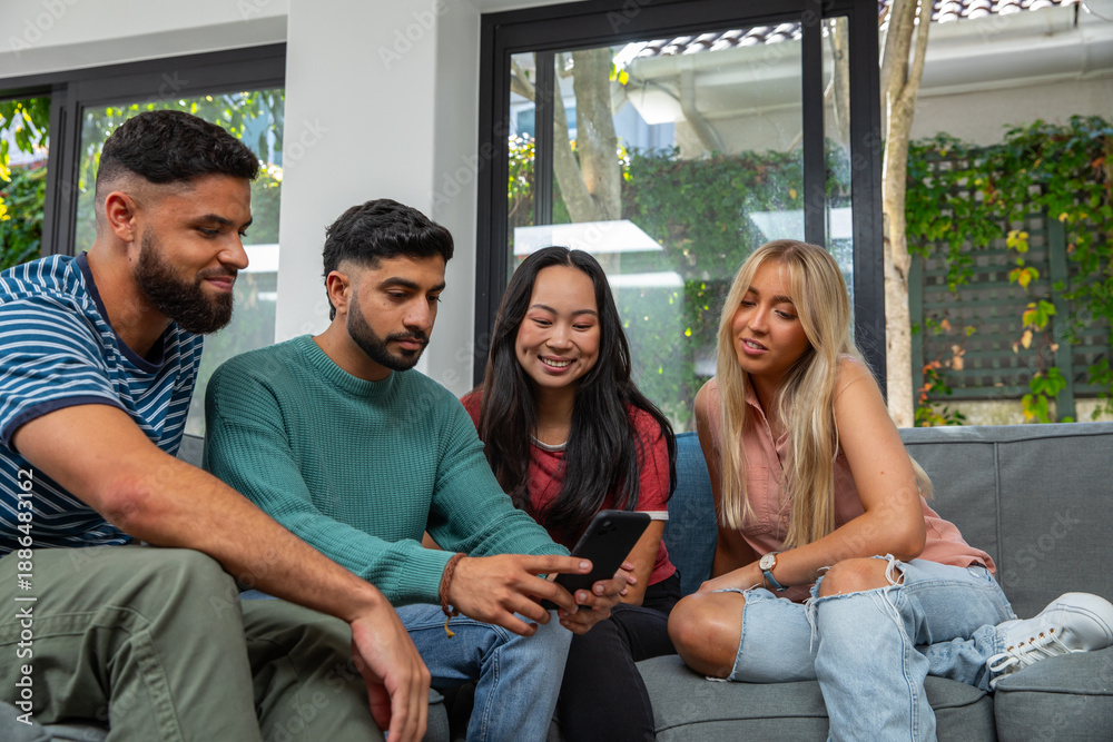 Fototapeta premium Diverse friends leaning in, watching smartphone on gray couch in bright living room