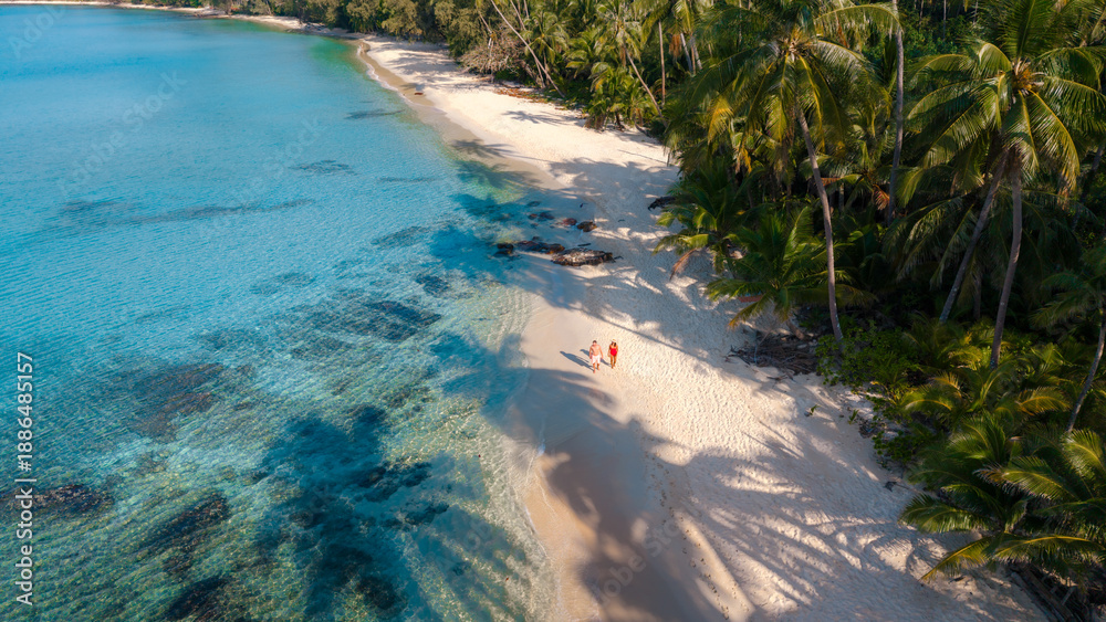 Fototapeta premium A couple experiences the tranquility of Takhian Beach on Koh Kood, Thailand, where two visitors stroll along the soft, powdery sands beneath tropical palm trees, embraced by the azure sea.