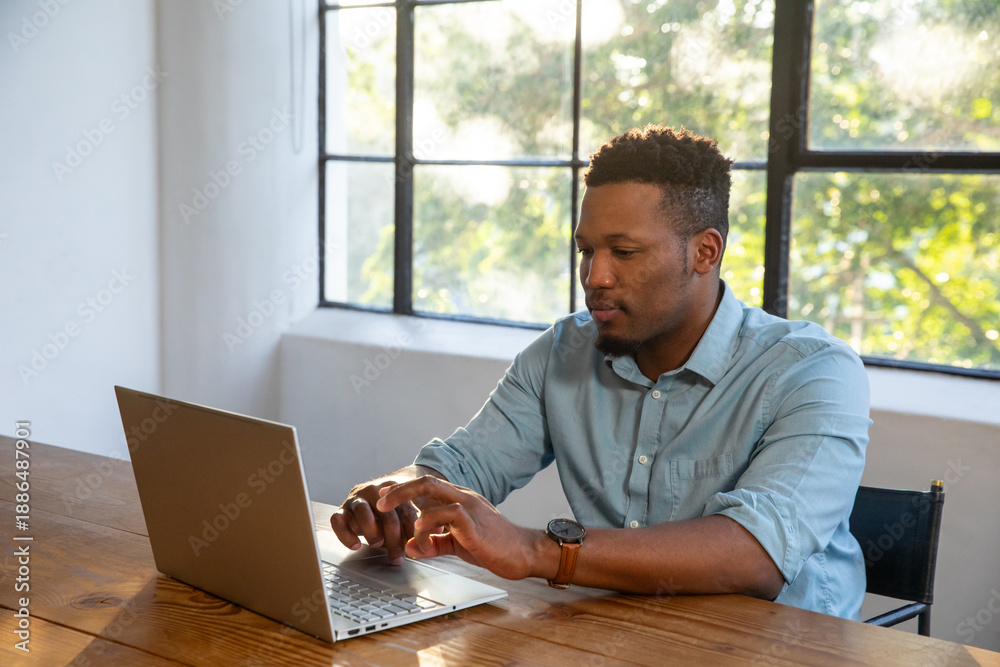 Fototapeta premium African american man typing on laptop at wooden table in office with foliage, copy space