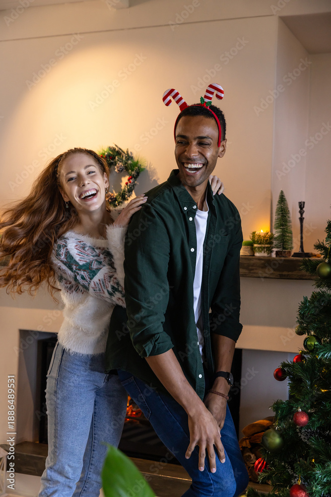 Naklejka premium Diverse couple dancing playfully in festive living room with reindeer headband, christmas tree