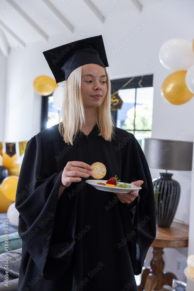 Naklejka premium Female graduate standing in living room wearing gown holding plate with crackers, strawberries