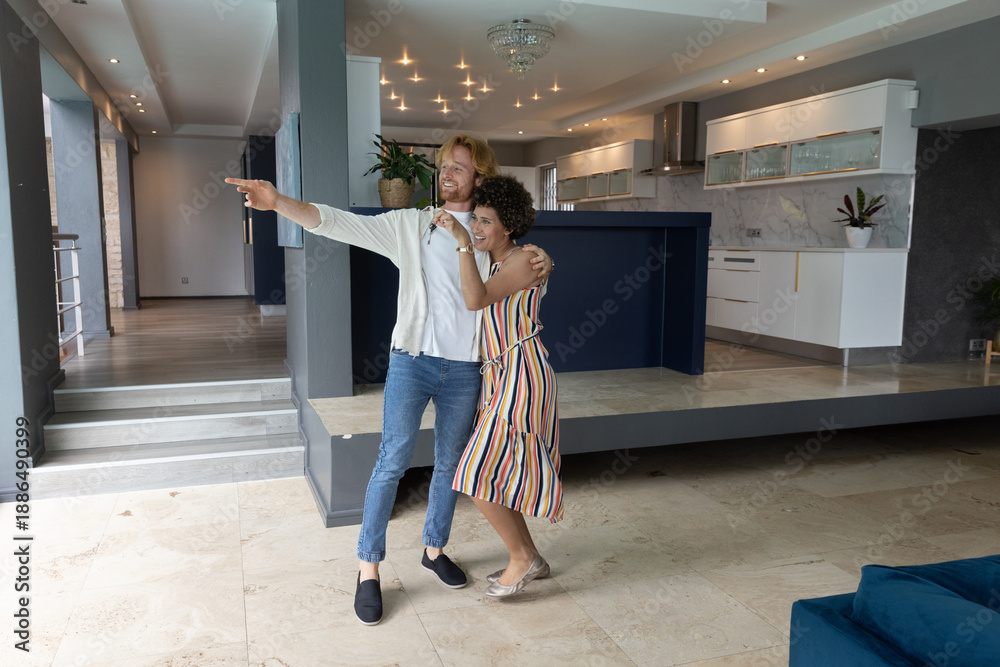 Naklejka premium Diverse couple standing in open-plan home area, embracing while pointing at kitchen island