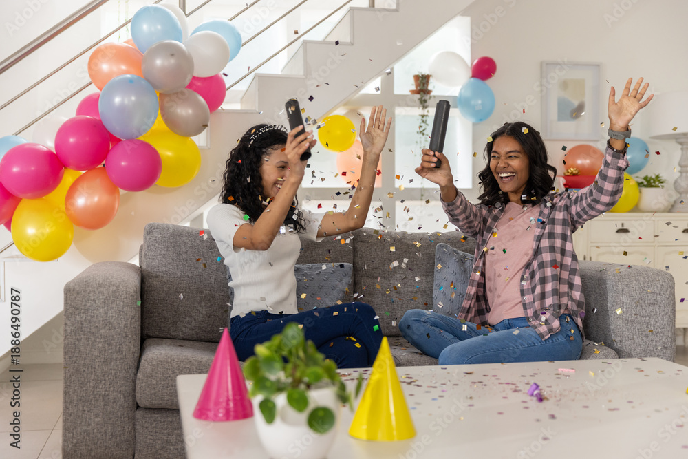 Obraz premium Diverse mom and teenage daughter sitting on sofa at home, popping black confetti poppers