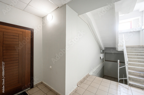 Foto Narrow stairwell with tiled floor, wooden door on left, white walls, ceiling light fixture, concrete stairs with white railing, small landing area