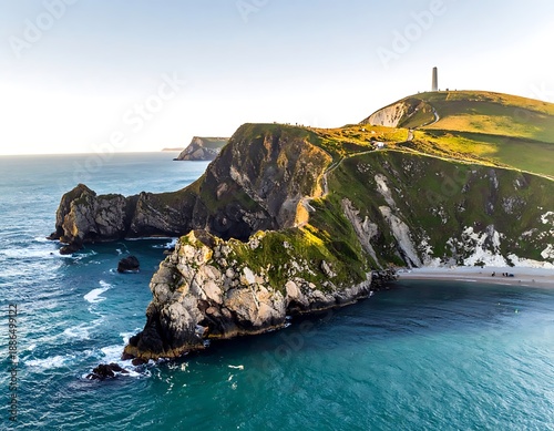 Aerial view of a grassy cliffside meeting the ocean, with a tower visible in the distance under a bright sky