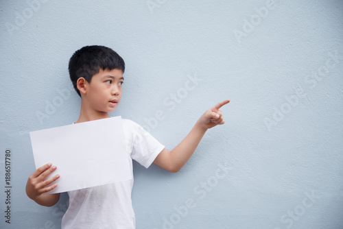 A young boy wearing a white shirt holds a white sheet of paper and points to a blank space on a gray background.