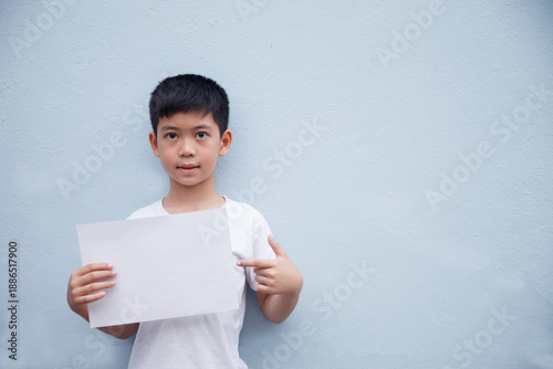 A boy wearing a white shirt holds a white piece of paper and points his finger at the white paper against a gray background.