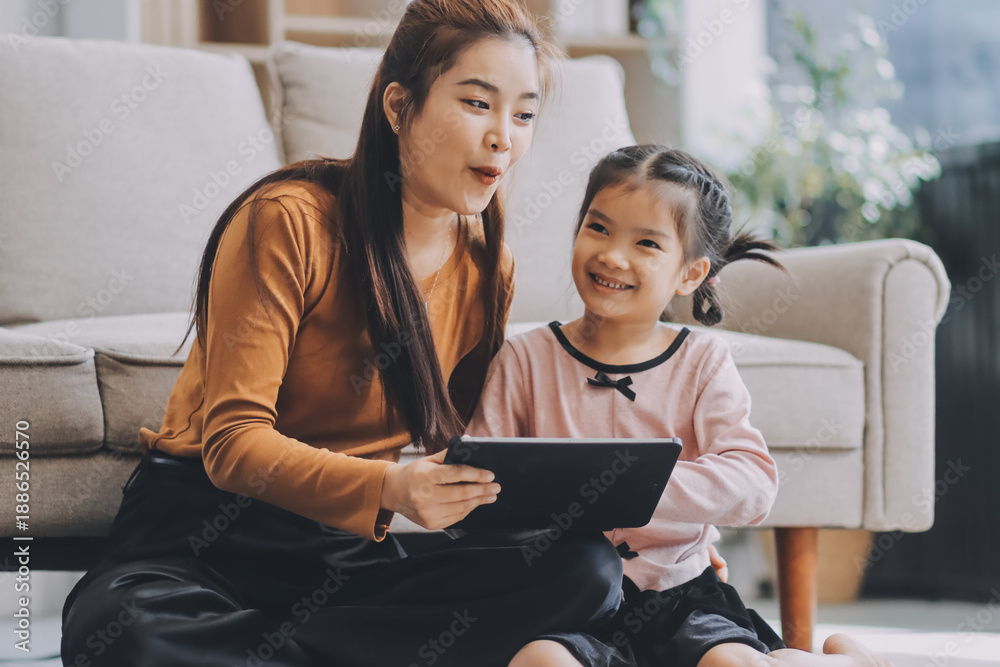 Fototapeta premium A cheerful Asian mother helps her young daughter do homework and color a book at home, while the father works on a laptop in the background. A cozy, bright, and modern family lifestyle.