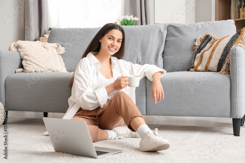 Pretty young woman with cup of hot coffee and laptop sitting on floor at home