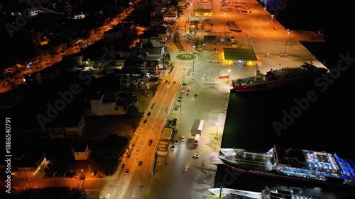 Night aerial drone view of the Port of Igoumenitsa, Greece. Flying over the busy ferry terminal with docked ships, trucks, and car traffic on the illuminated coastal road