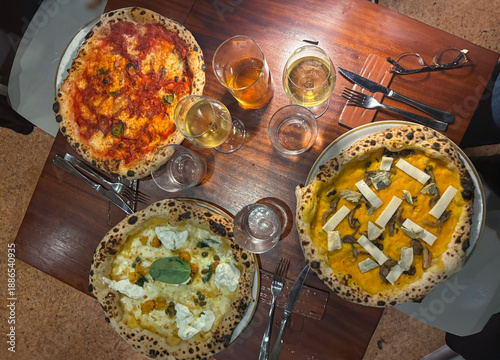 Top view of three delicious gourmet Neapolitan pizzas with leopard crust on a wooden table, served with white wine and beer in a cozy restaurant atmosphere, authentic Italian dinner party.