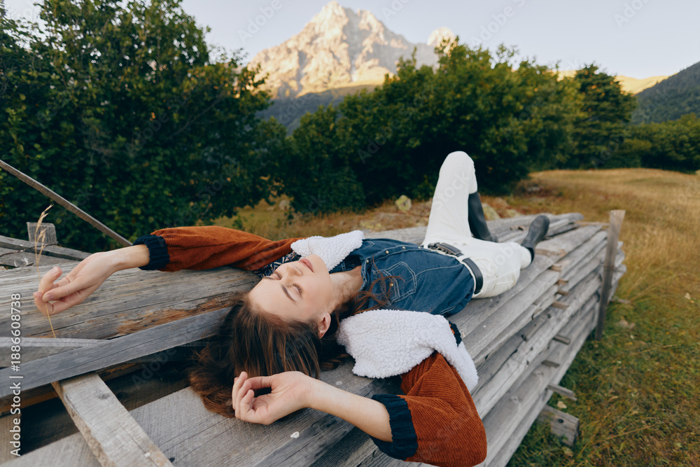 Fototapeta premium Woman relaxation on wooden platform in countryside meadow with mountain and nature views, lying outdoors in denim jacket and shearling, peaceful summer rest and scenic escape.