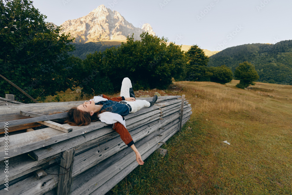 Fototapeta premium Woman lying on a wooden fence in a meadow with a mountain view, relaxing in nature among trees and open countryside, wearing casual jacket for a peaceful outdoor escape.