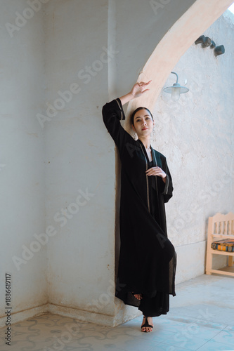 A full-length woman in a black abaya against a street background in soft natural light. Traditional Arab-Muslim clothing.