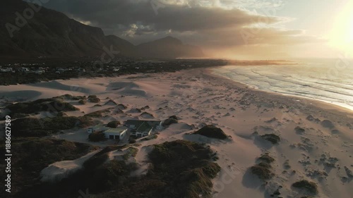 Aerial view of sandy beach meeting the ocean, with mountains looming in the background under a dramatic sky filled with clouds, Betty's Bay, Western Cape, South Africa.
