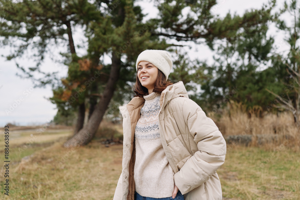 Obraz premium Woman in a warm beige coat and cream sweater stands in a windy park, wearing a knit beanie, casual winter fashion with trees and open field in the background.