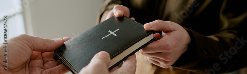 Website banner of Caucasian adult hands giving Bible with silver cross to Caucasian child hands, close up showing intergenerational religious connection and faith sharing