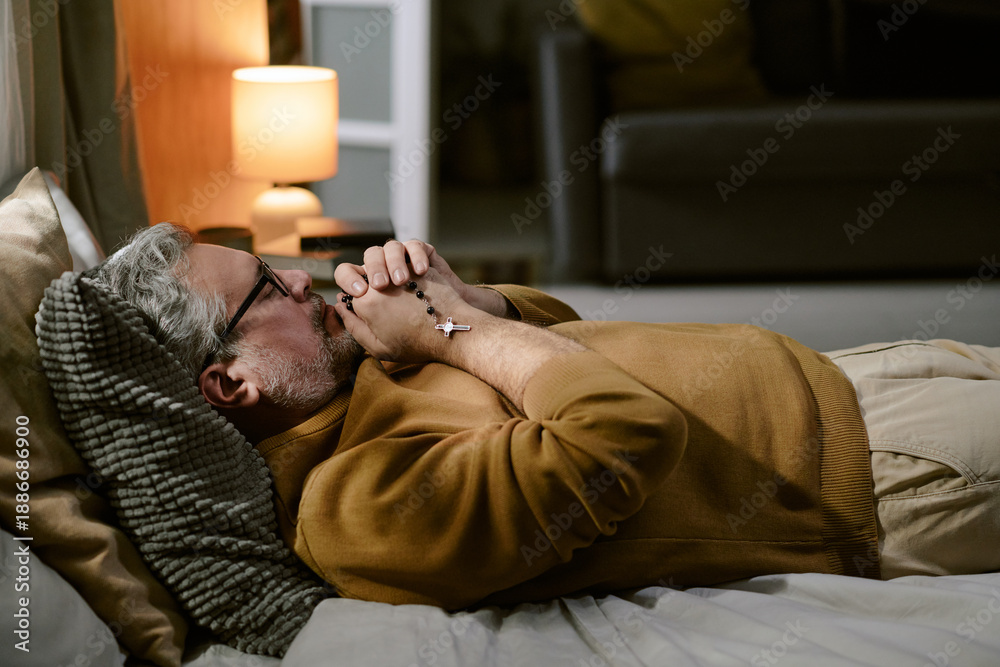 Obraz premium Middle aged Caucasian man lying on bed holding rosary beads with clasped hands near face appearing to pray or meditate, wearing glasses, relaxing in softly lit bedroom