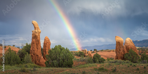 View of vibrant rainbow arcs across the sky, illuminating the textured sandstone formations and green vegetation in a striking contrast, Escalante, Utah, United States.