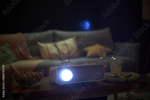 Home setting showing digital projector turned on with bright lens light, surrounded by bowls of popcorn, snacks, and drinks on wooden table in front of sofa