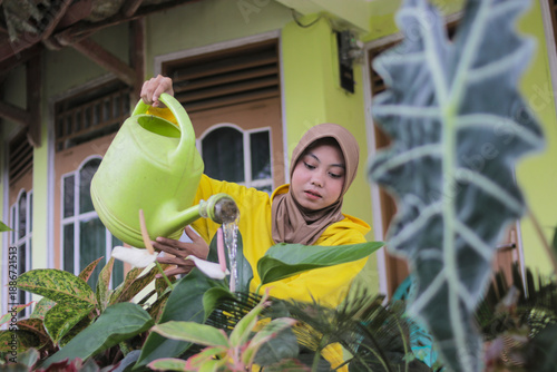 Fotografie A young Muslim woman wearing a hijab and a casual yellow hoodie waters potted pl