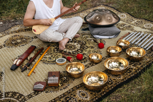 Wide view of a musician seated outdoors surrounded by sound healing instruments, including handpan, singing bowls and flutes, creating a serene wellness atmosphere.