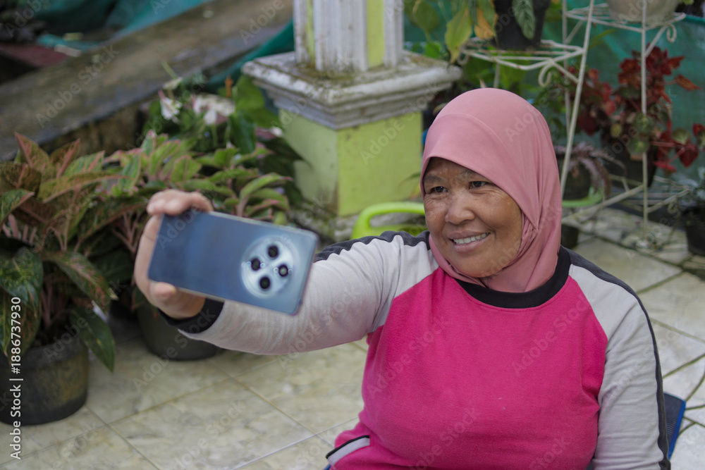 Naklejka premium Middle-aged Asian woman wearing a hijab takes a selfie on her home terrace after completing a workout. Fitness, healthy lifestyle, and home exercise concept.