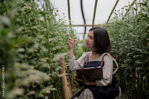 Fotografija Young Asian woman florist with short hair wearing a gardening apron writes notes on a clipboard while checking flower growth inside a greenhouse