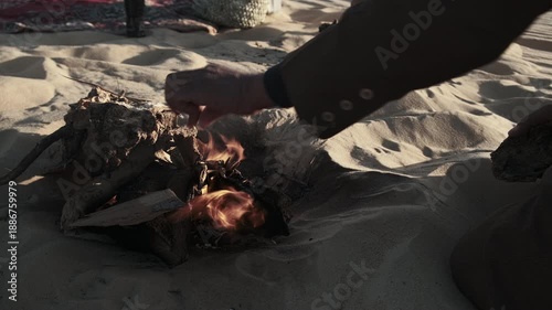 Hand of man dressed in traditional berber clothes tending small open fire in desert sand. Nomadic lifestyle, cultural ritual, survival practice in arid environment. Sahara desert, Siwa oasis, Egypt