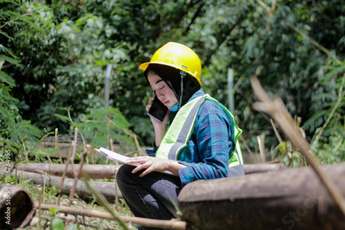 Asian woman wearing a yellow hard hat and safety vest talks on her phone while writing notes on a clipboard in a forest. Field inspection, environmental work, and professional documentation concept