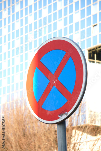 Fotografie Close-up of a red and blue no stopping traffic sign against a blurred modern building and tree branches