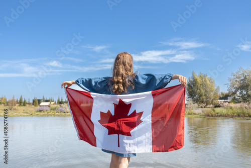Happy Canada Day. An unrecognizable young woman holds a large Canadian flag behind her on a sunny day. Travel, immigration. Pride, celebration, national symbol.